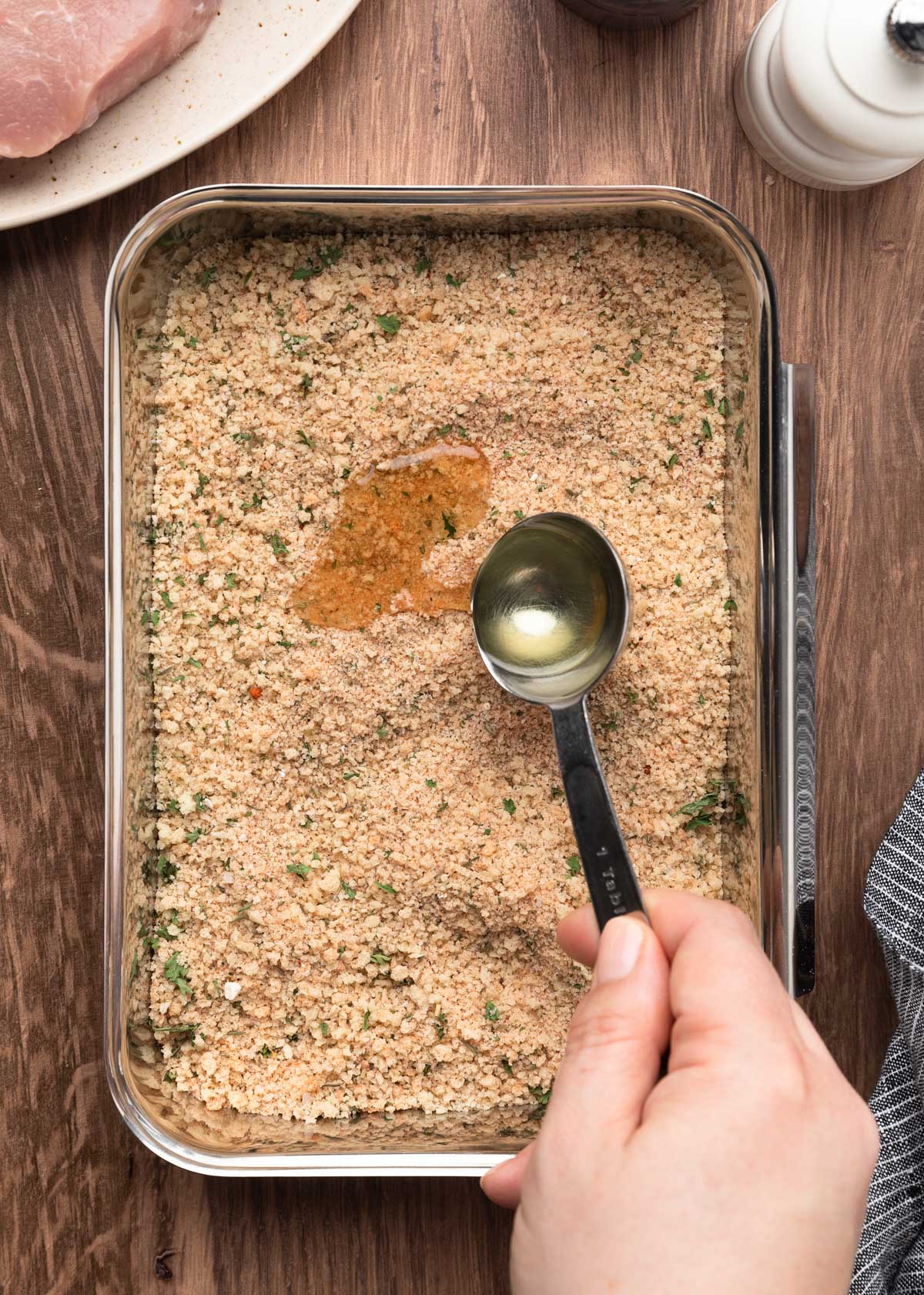 Hand pouring oil from a tablespoon over a seasoned breadcrumb mixture in a metal pan, preparing the breading for pork chops.