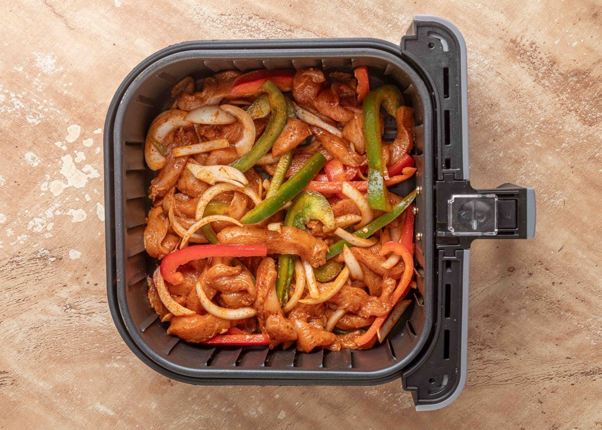 Seasoned raw chicken strips, bell peppers, and onions arranged in a single layer in an air fryer basket, ready to cook for fajitas.