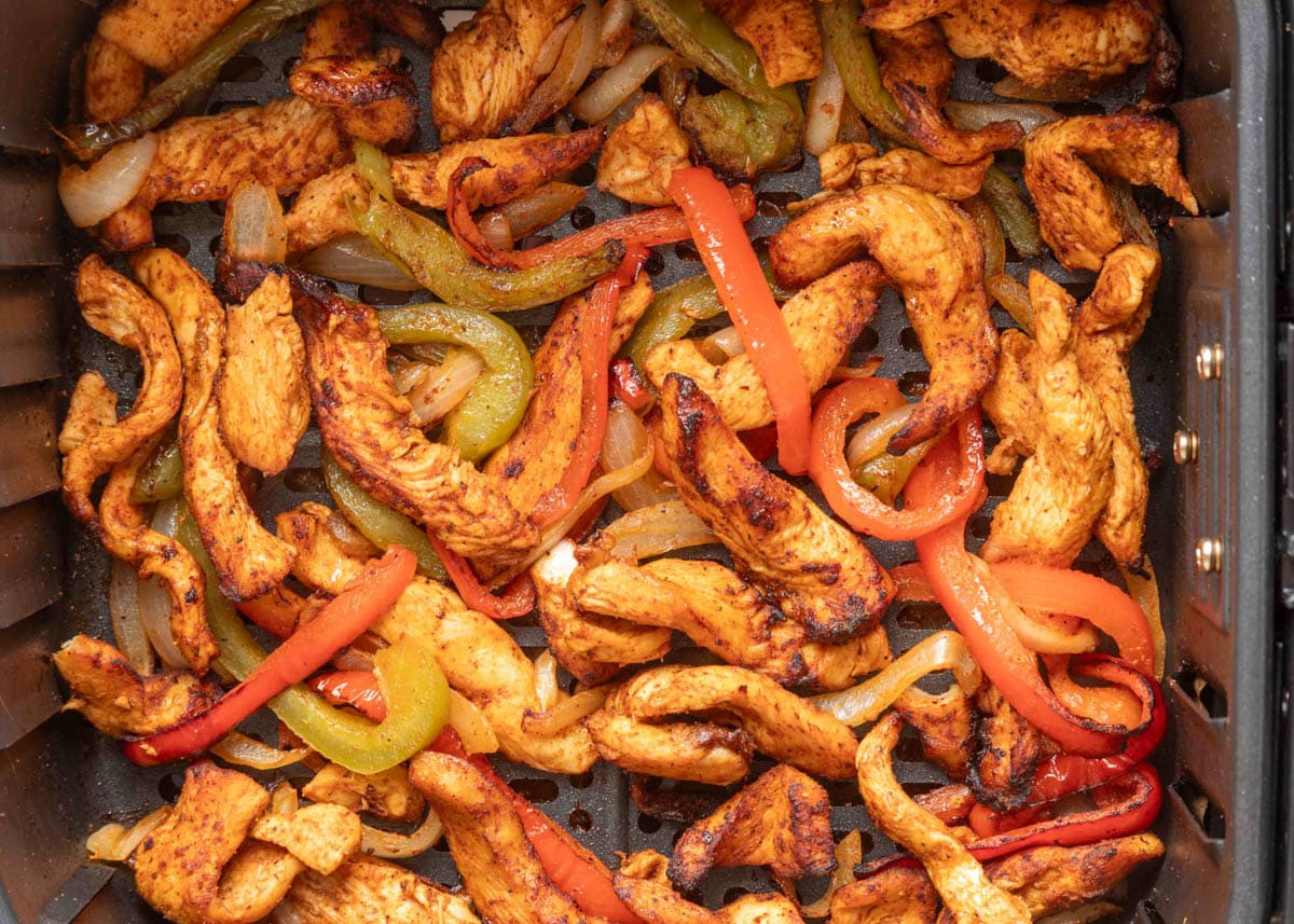 Cooked chicken fajita strips, red and green bell peppers, and onions in an air fryer basket, lightly charred and ready to serve.