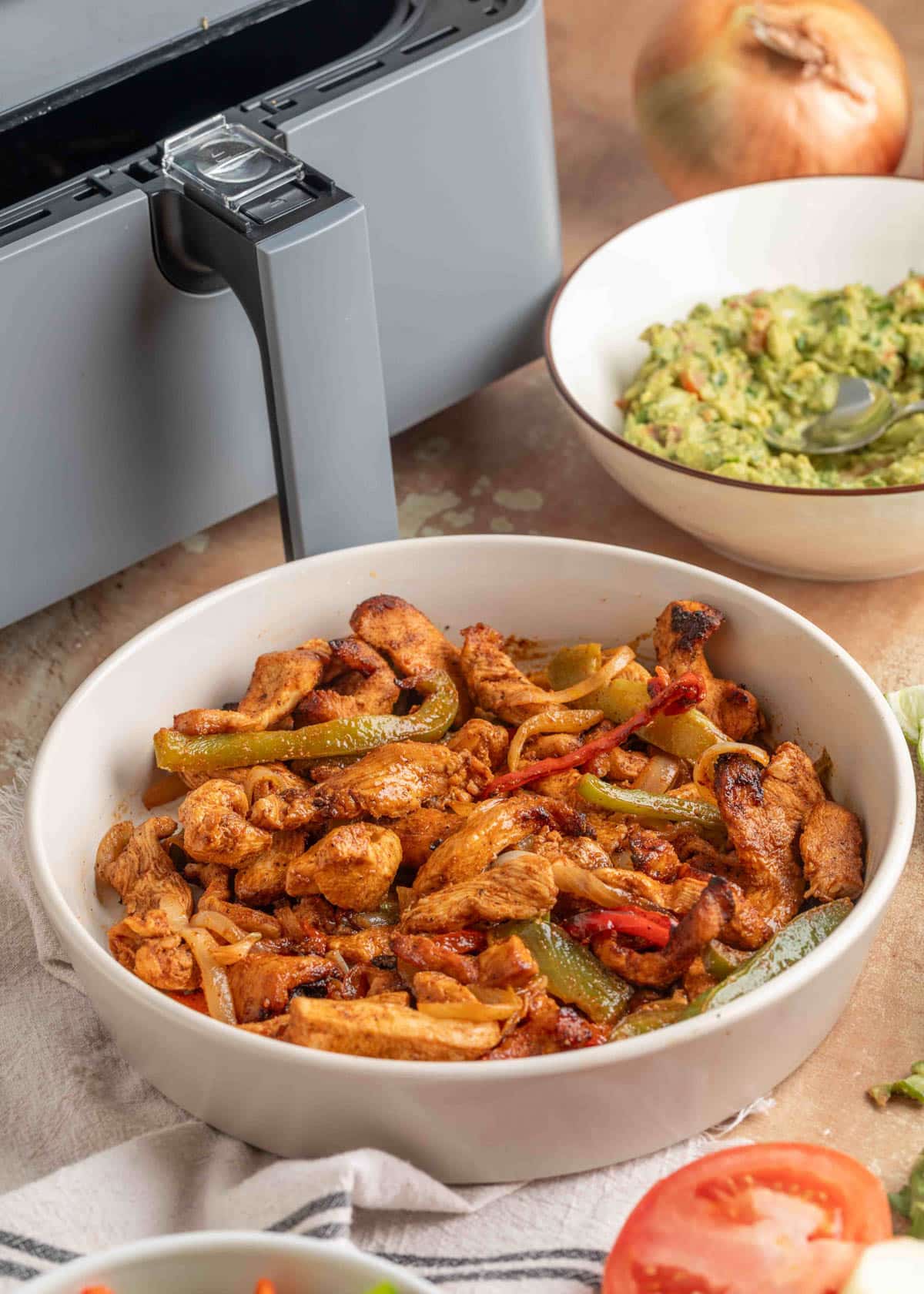 Bowl of air fryer chicken fajitas with saut&eacute;ed peppers and onions, set on a cloth next to an air fryer and a bowl of guacamole.