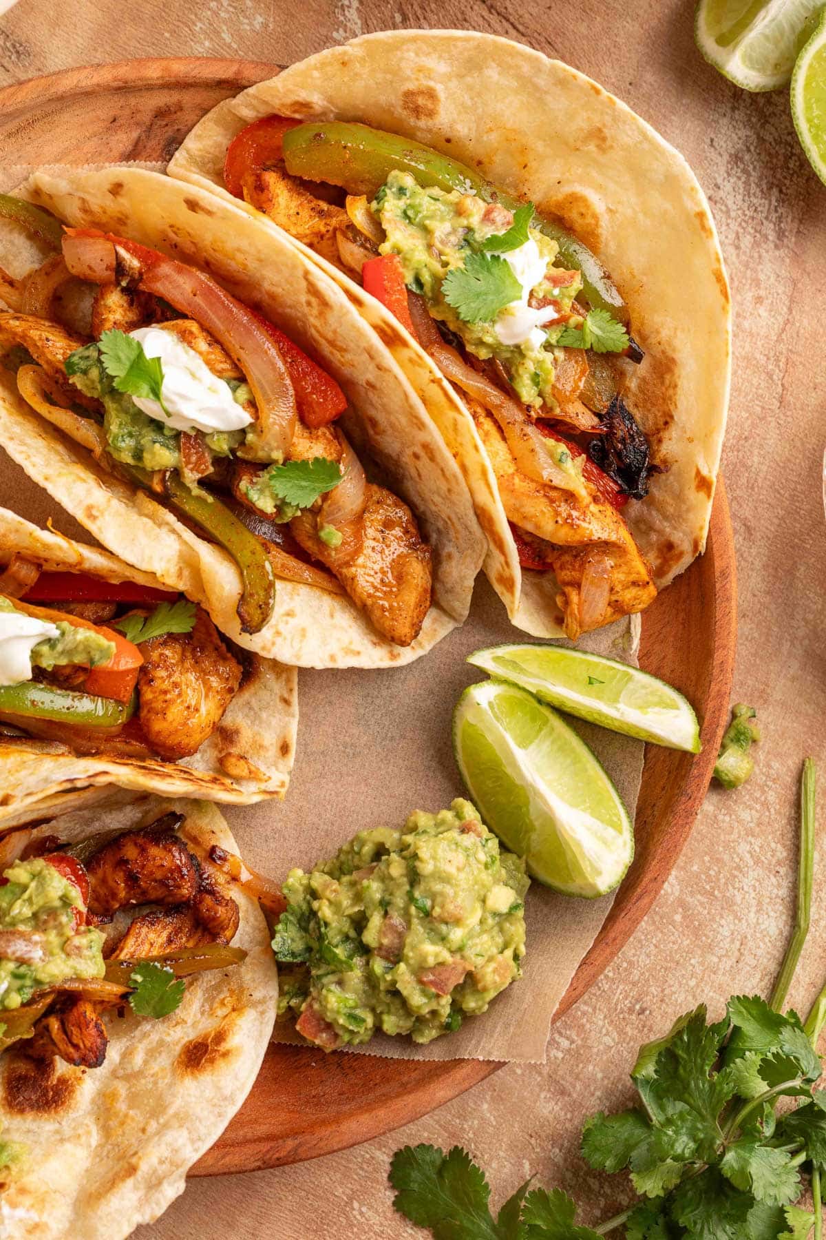 Plated air fryer chicken fajita tacos in flour tortillas topped with guacamole, sour cream, and cilantro, served with lime wedges and extra guacamole on a wooden board.