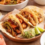 Four air fryer chicken fajita tacos in flour tortillas filled with seasoned chicken, sautéed peppers, and onions, served on a wooden plate with guacamole and lime wedges.