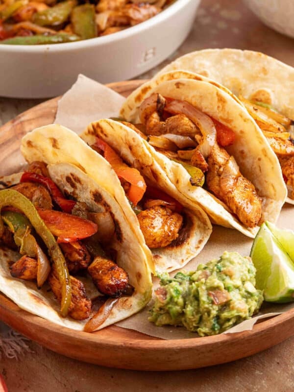 Four air fryer chicken fajita tacos in flour tortillas filled with seasoned chicken, saut&eacute;ed peppers, and onions, served on a wooden plate with guacamole and lime wedges.