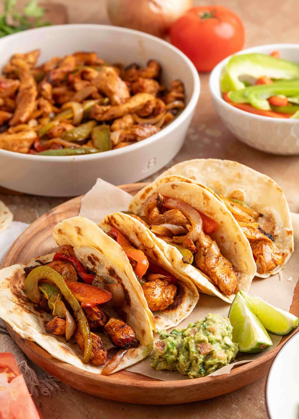 Four flour tortillas filled with air fryer chicken fajitas, including seasoned chicken strips, saut&eacute;ed onions, and bell peppers, served on a wooden plate with lime wedges and a scoop of chunky guacamole. A bowl of extra fajita filling and sliced peppers are visible in the background.