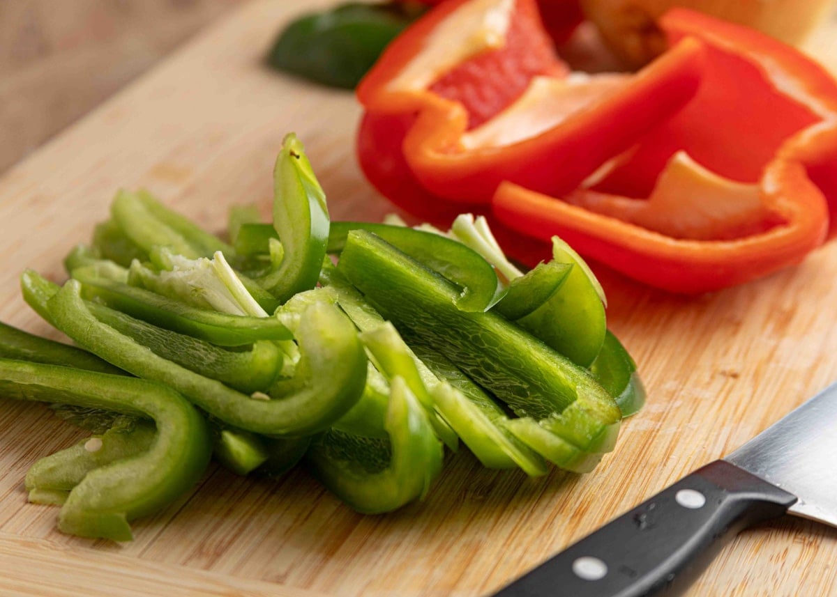 Freshly sliced green bell peppers on a wooden cutting board with halved red peppers in the background and a paring knife nearby.