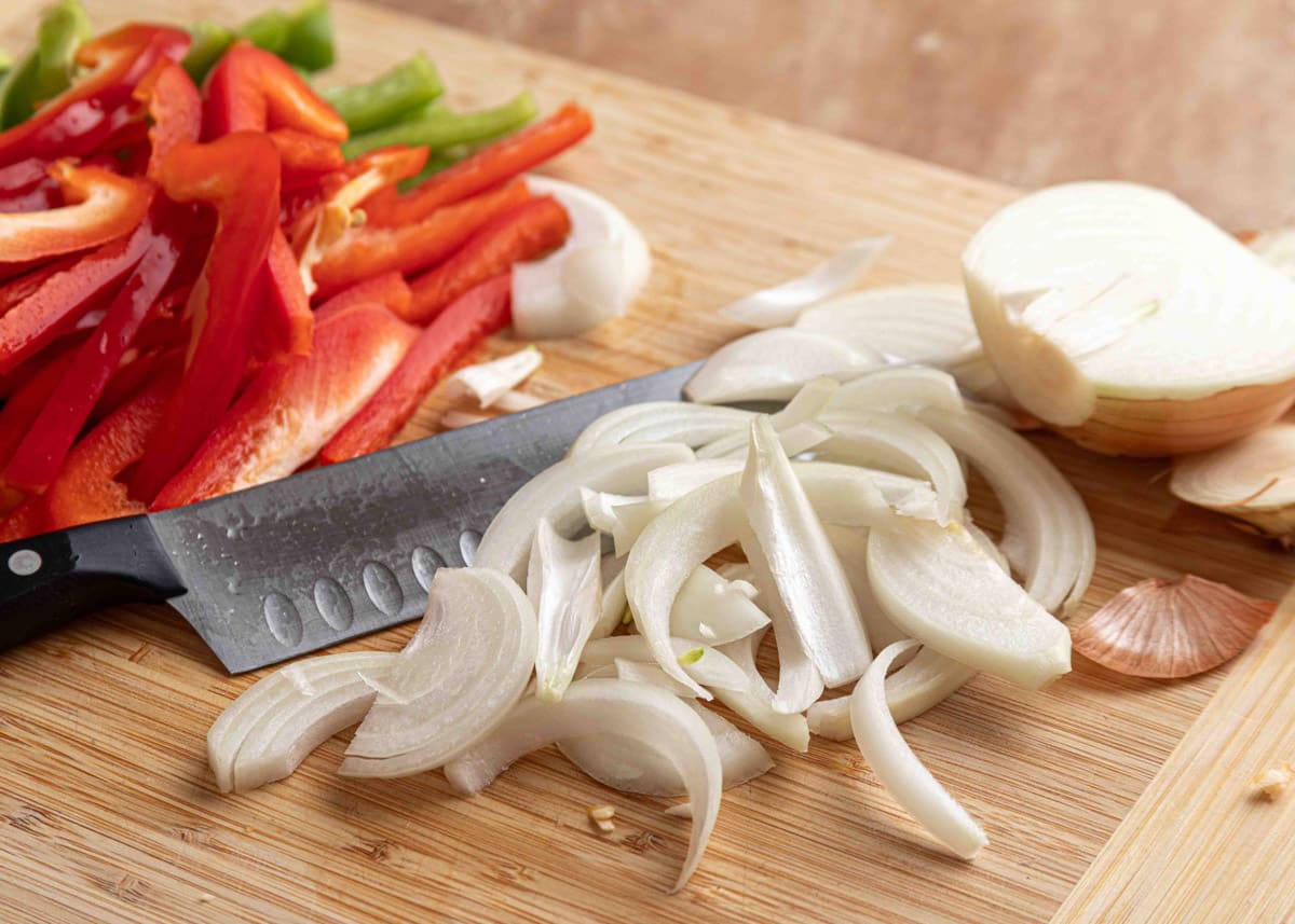 Sliced yellow onions and red bell peppers on a wooden cutting board with a chef&rsquo;s knife, ready to be cooked for chicken fajitas.