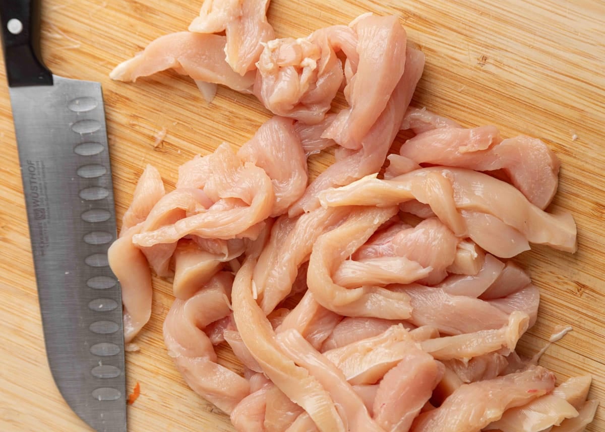 Raw chicken breasts sliced into thin strips on a wooden cutting board with a chef&rsquo;s knife, ready for fajita preparation.