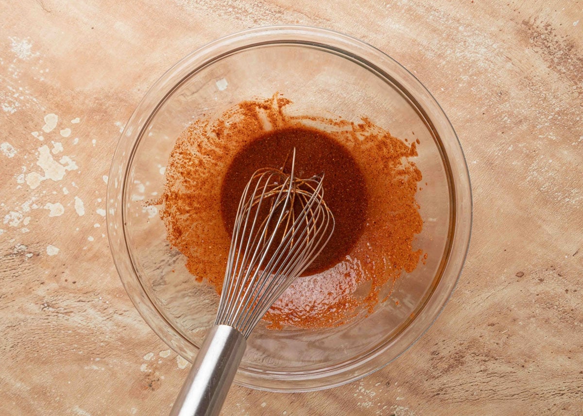 A stainless steel whisk in a glass bowl filled with a freshly mixed reddish-brown fajita marinade on a tan textured surface.