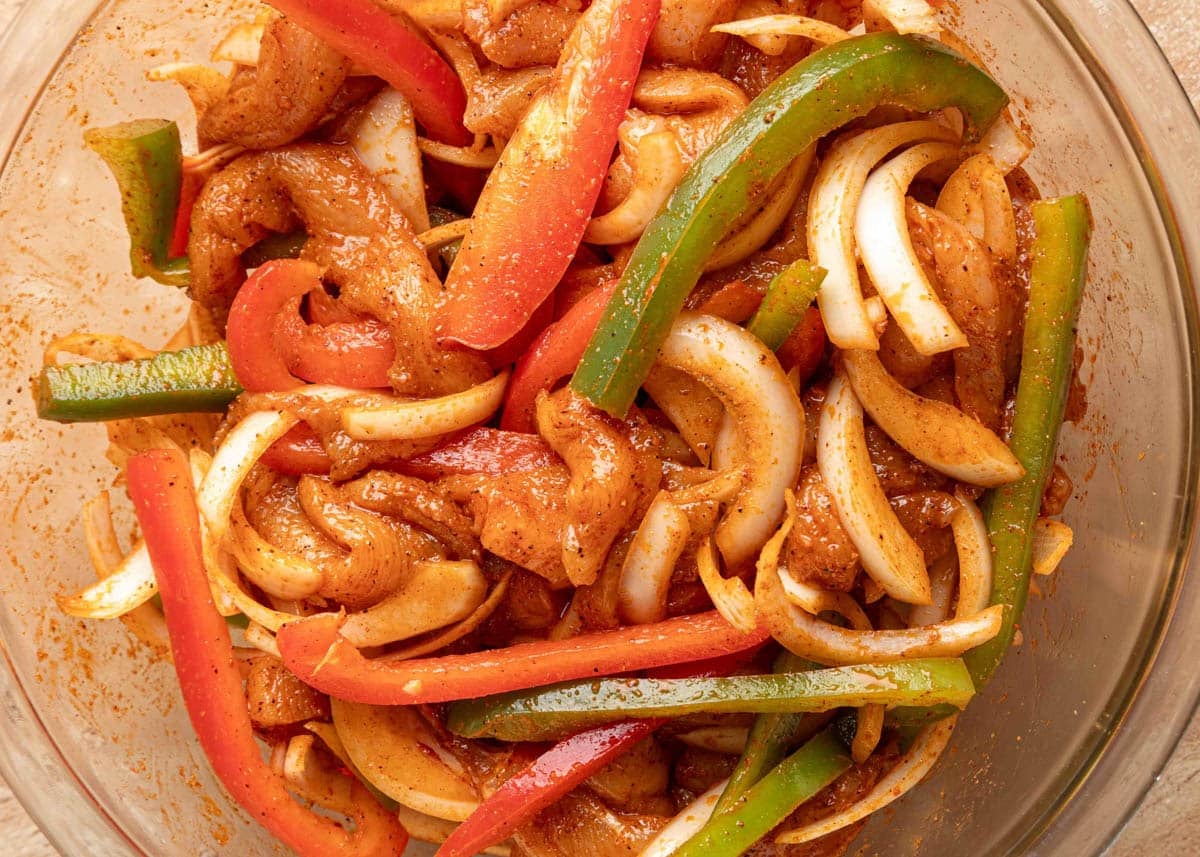 Close-up of sliced raw chicken, red and green bell peppers, and onions coated in a seasoned marinade in a glass mixing bowl, ready to cook for fajitas.