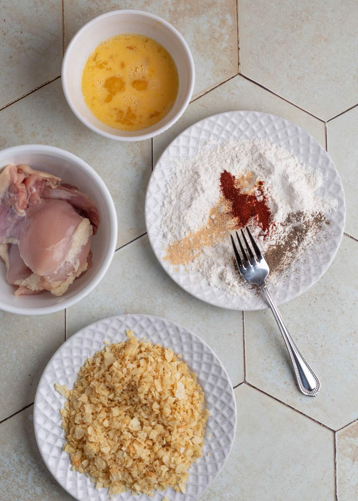 Overhead view of prep bowls with raw chicken, egg wash, crushed potato chips, and a white plate of flour with paprika, garlic powder, onion powder, pepper, and a fork ready to mix.