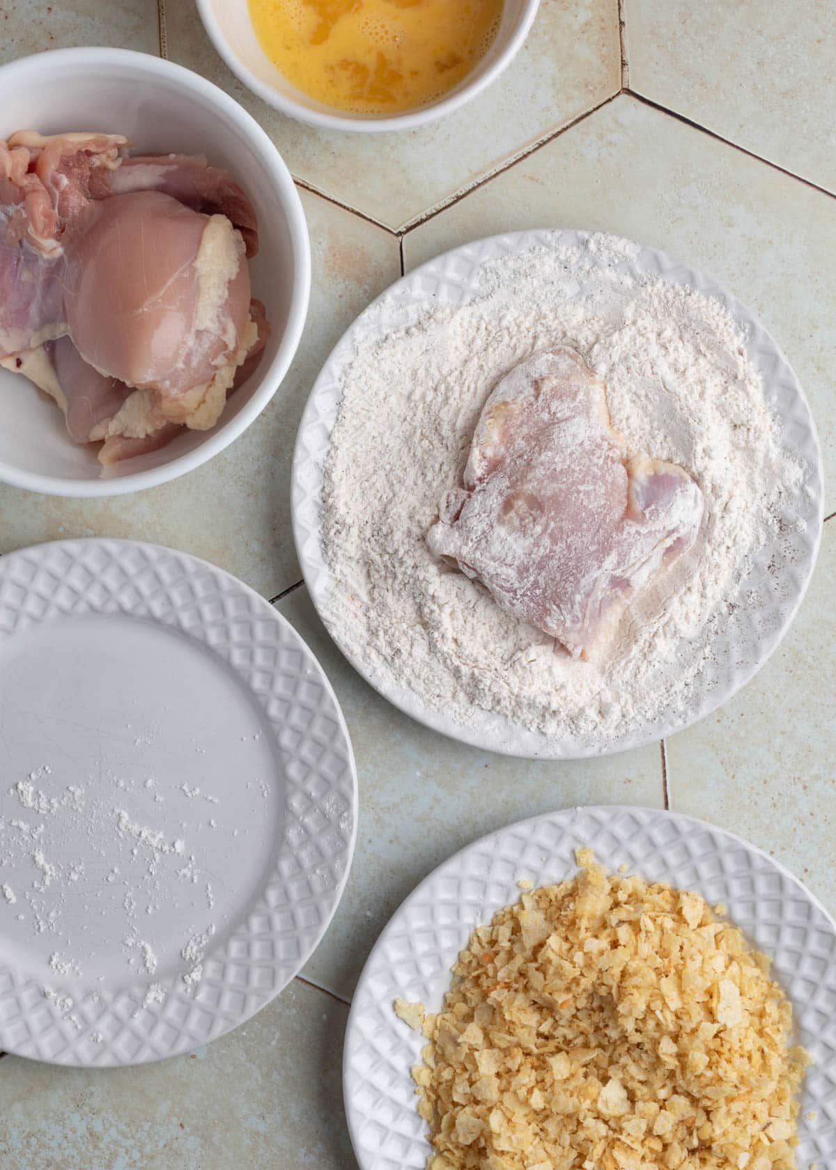Raw chicken thigh dredged in seasoned flour, resting on a white plate, next to bowls of egg wash, raw chicken thighs, and crushed chips.