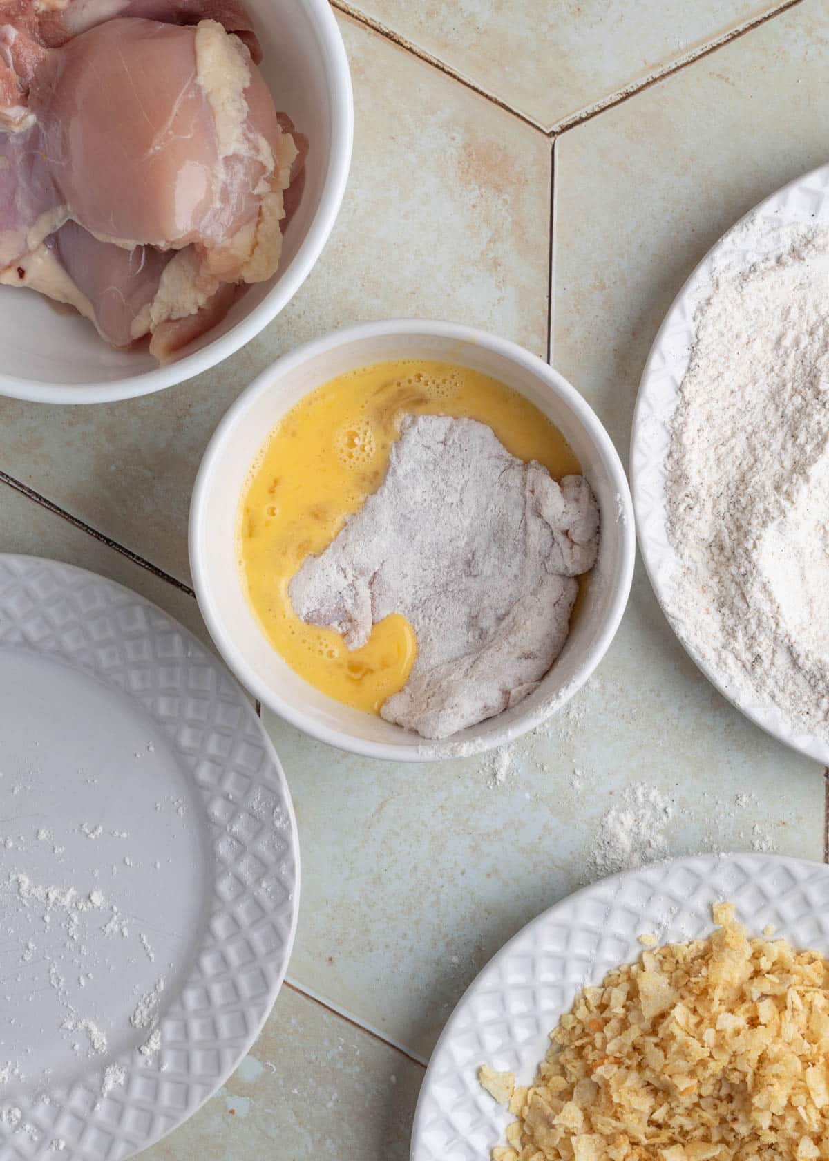 Flour-dredged raw chicken thigh being dipped into a bowl of beaten eggs, with surrounding bowls of raw chicken, seasoned flour, and crushed potato chips.