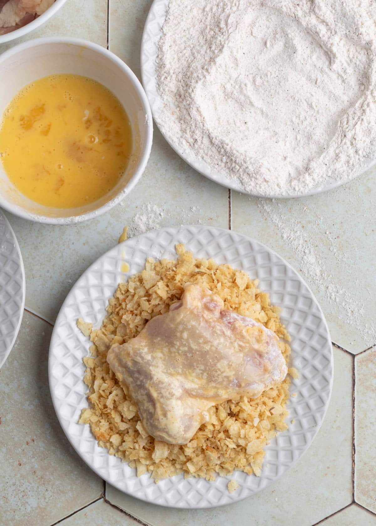 Raw chicken thigh coated in egg wash, resting on a plate of crushed potato chips, surrounded by bowls of flour and beaten eggs on a tiled countertop.