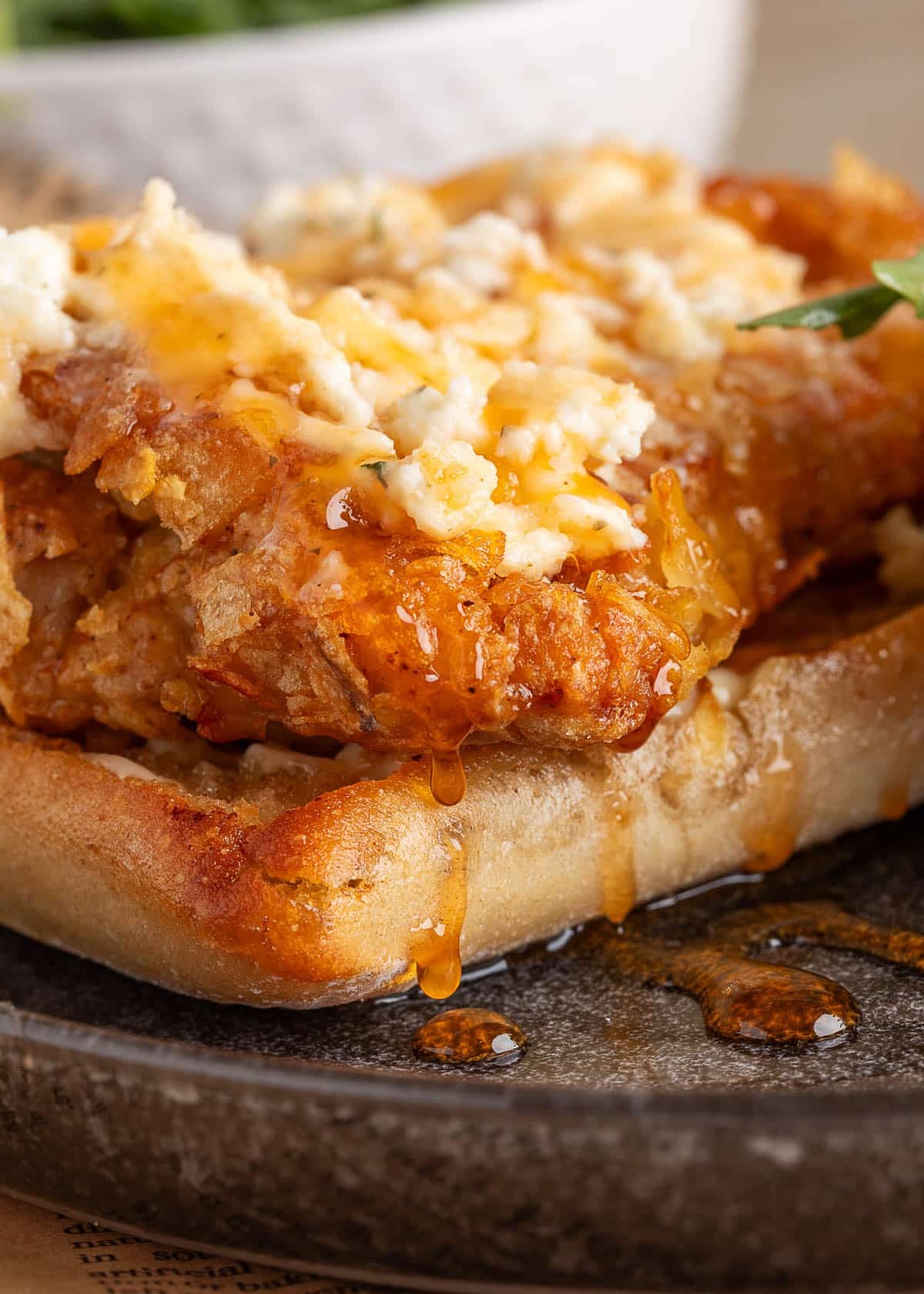 Macro shot of the sandwich&rsquo;s crispy chicken topped with Boursin cheese and a thick drizzle of golden honey pooling onto the plate, showcasing rich textures and shine.