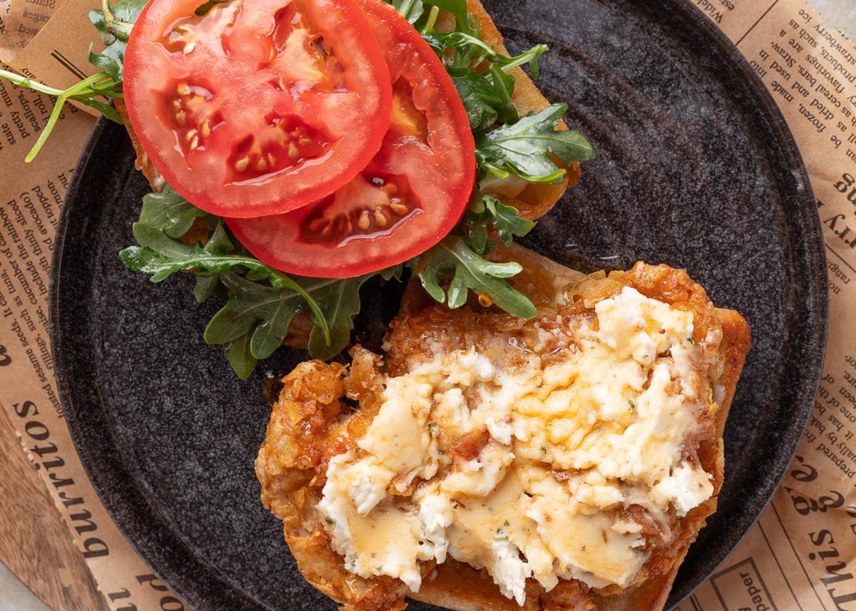 Overhead view of an open-faced air fryer chicken sandwich with potato chip-crusted chicken topped with Boursin and hot honey on one side, and fresh arugula and tomato slices on the other, served on a toasted ciabatta roll.
