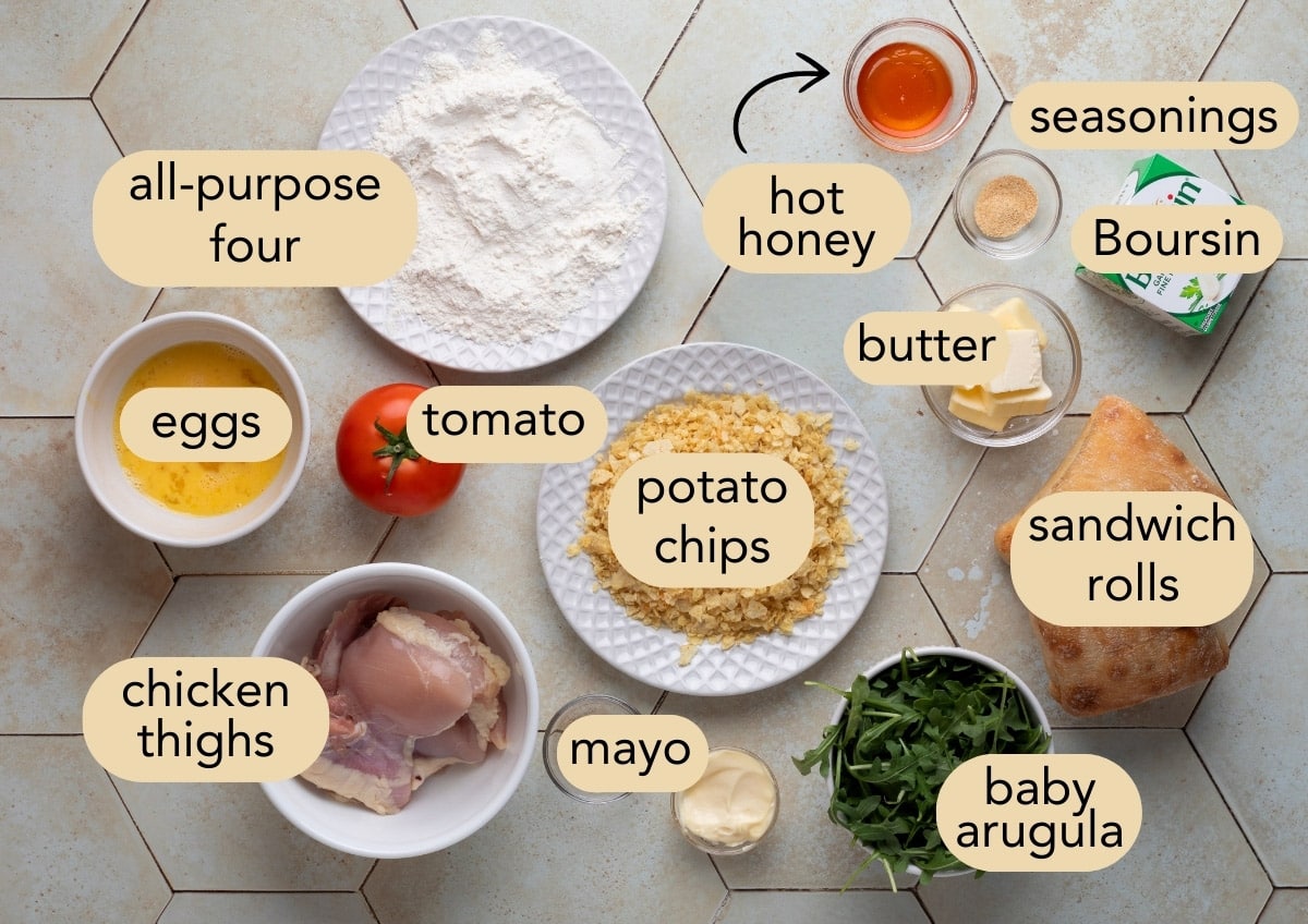 Flat lay of labeled ingredients for a crispy chicken sandwich with hot honey, including chicken thighs, crushed potato chips, sandwich rolls, Boursin cheese, baby arugula, tomato, mayo, eggs, flour, butter, hot honey, and seasonings on a tiled surface.