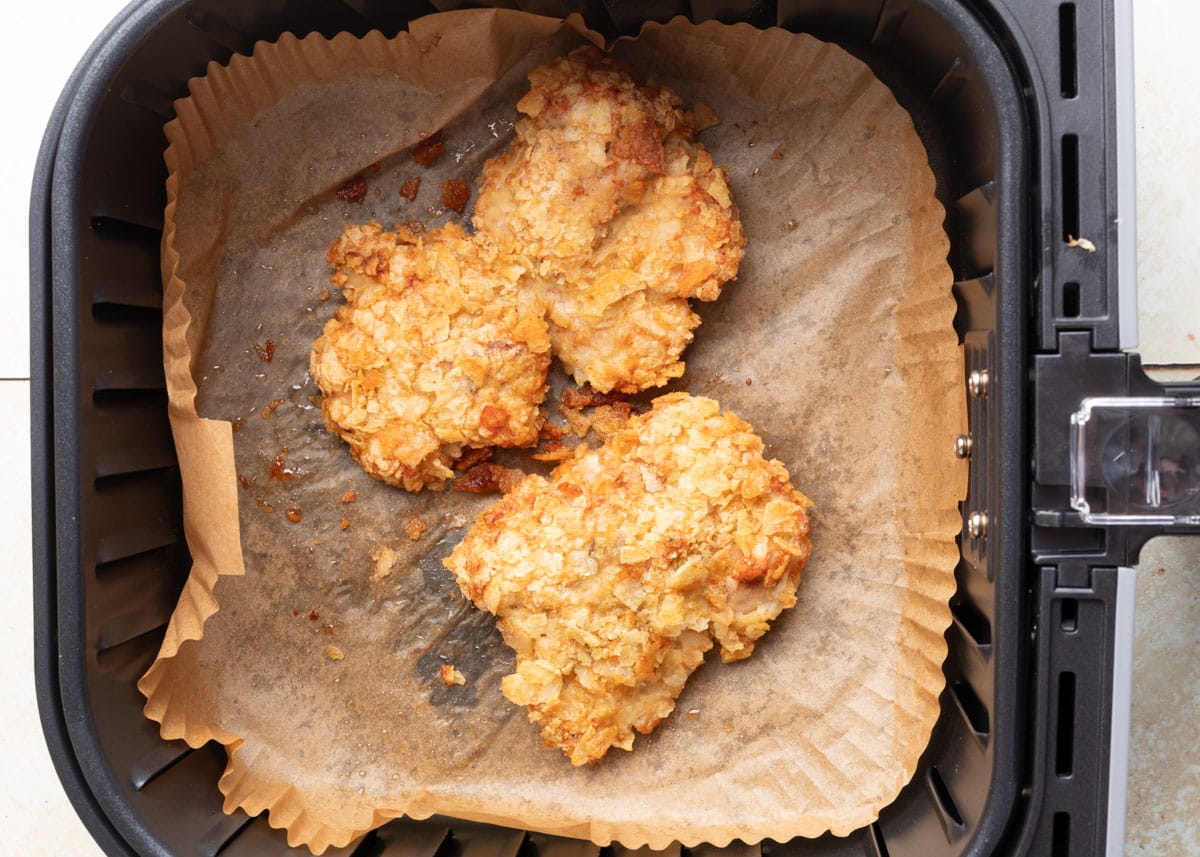 Two crispy potato chip&ndash;crusted chicken thighs in an air fryer basket lined with parchment paper, freshly cooked and golden brown.
