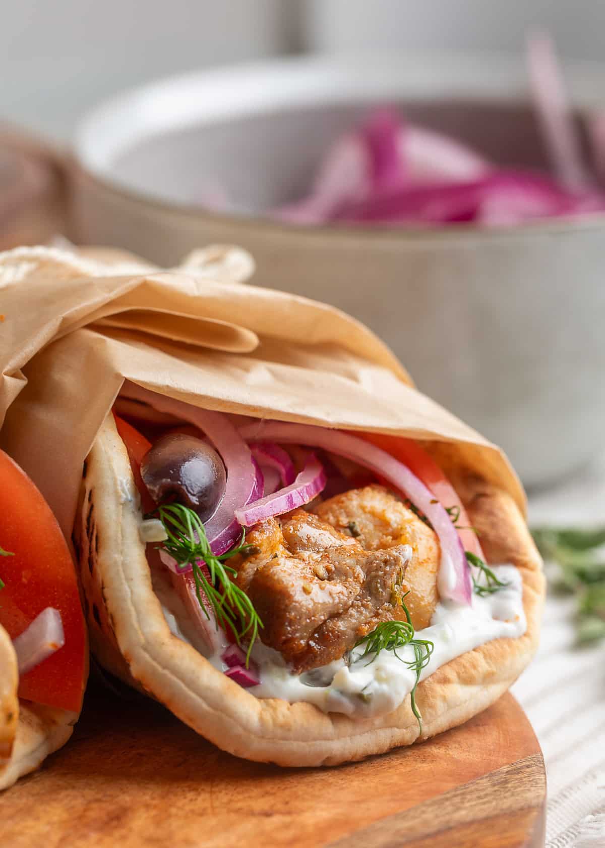 A close-up of a pita wrap filled with grilled chicken souvlaki, creamy tzatziki sauce, sliced red onions, fresh dill, tomato, and a Kalamata olive, wrapped in parchment paper and set on a wooden board. A bowl of red onions is softly blurred in the background.