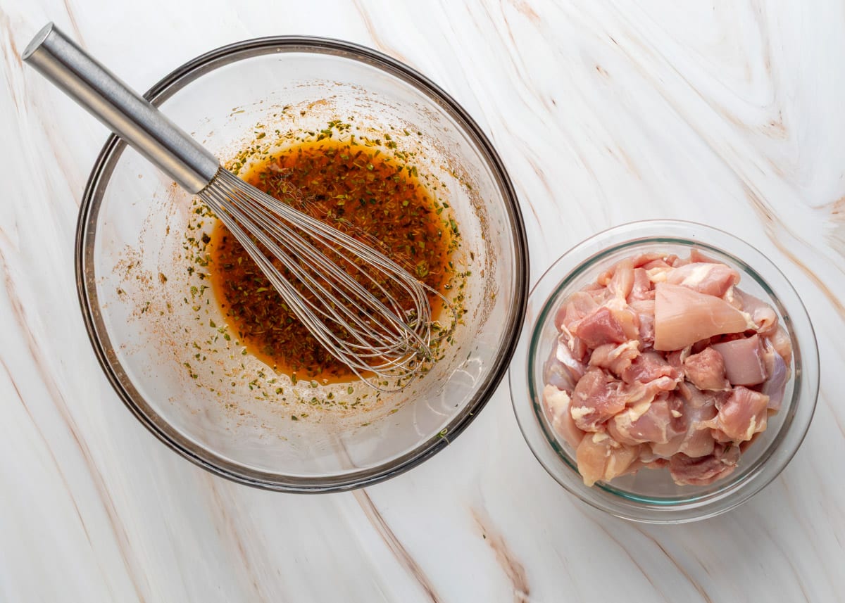 Overhead view of a glass mixing bowl filled with a red-tinged Greek-style marinade, with herbs visible throughout and a metal whisk resting inside. Below the bowl is a separate glass dish containing raw, cubed boneless chicken thighs, placed on a light marble surface.