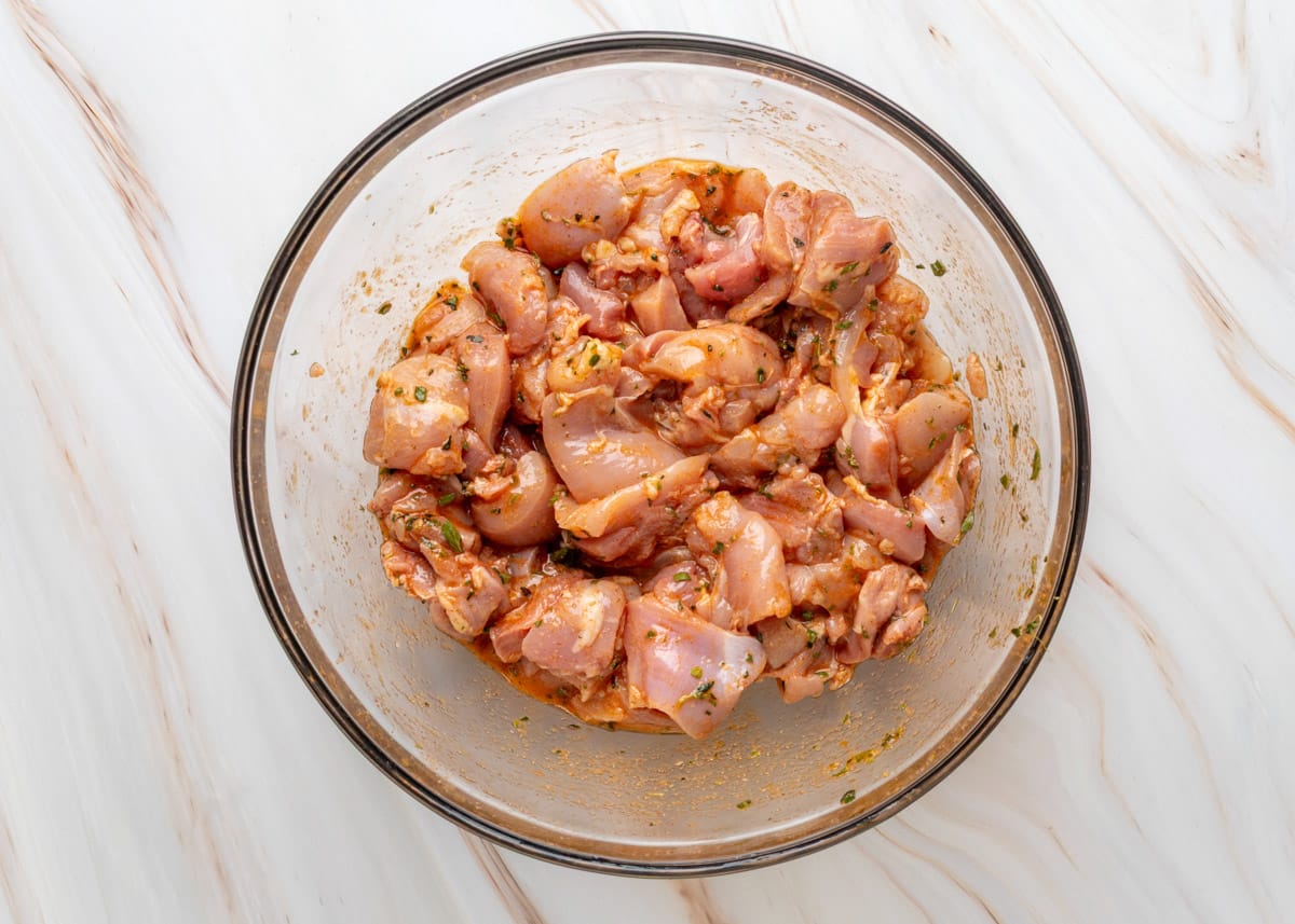 Marinated chicken thighs in a glass bowl, fully coated in a red-hued herb marinade. The chicken is cut into chunks and ready to be skewered or cooked, set against a white marble background.