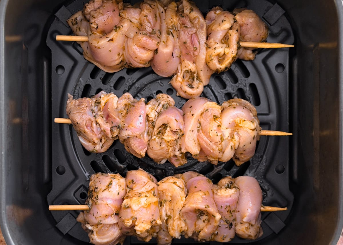Three skewers of marinated raw chicken thighs arranged in the basket of an air fryer, ready to cook, with visible herbs and seasoning coating the meat.