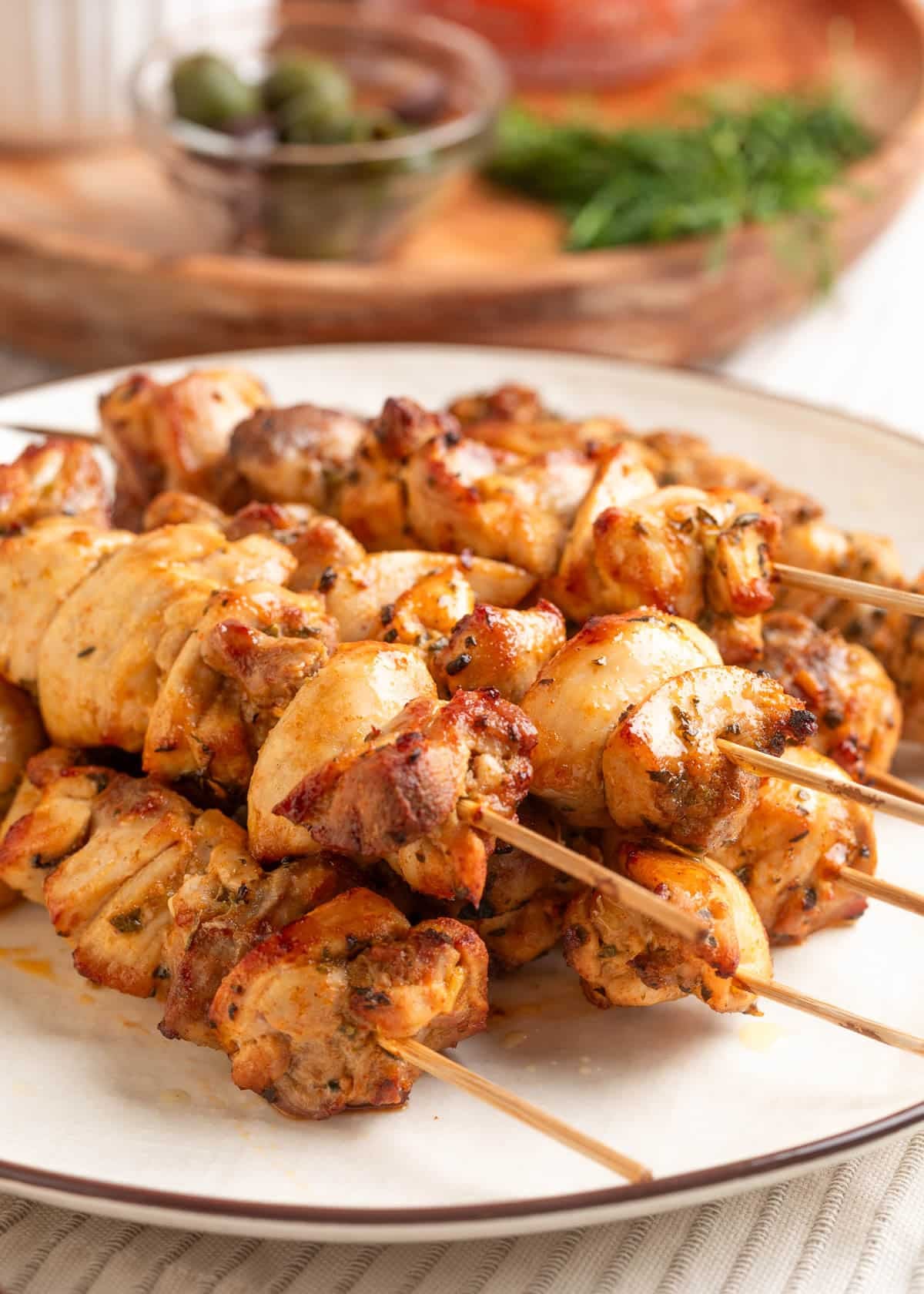 Close-up of juicy, golden-brown chicken souvlaki skewers stacked on a white plate, showing charred edges and herbs, with a small bowl of olives and fresh herbs softly blurred in the background.