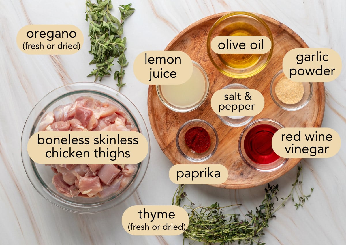 Overhead view of labeled ingredients for chicken souvlaki, including boneless skinless chicken thighs in a glass bowl, fresh oregano and thyme, and small bowls of lemon juice, olive oil, red wine vinegar, garlic powder, paprika, and salt and pepper arranged on a wooden tray.