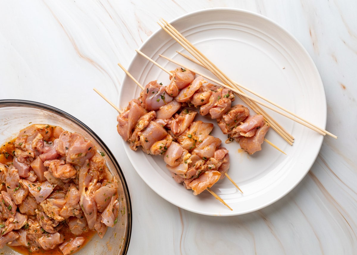 Overhead view of raw, marinated chicken thigh pieces threaded onto wooden skewers and arranged on a white plate. Additional skewers rest beside them, with the remaining marinated chicken visible in a large glass mixing bowl to the left, all set on a light marble surface.