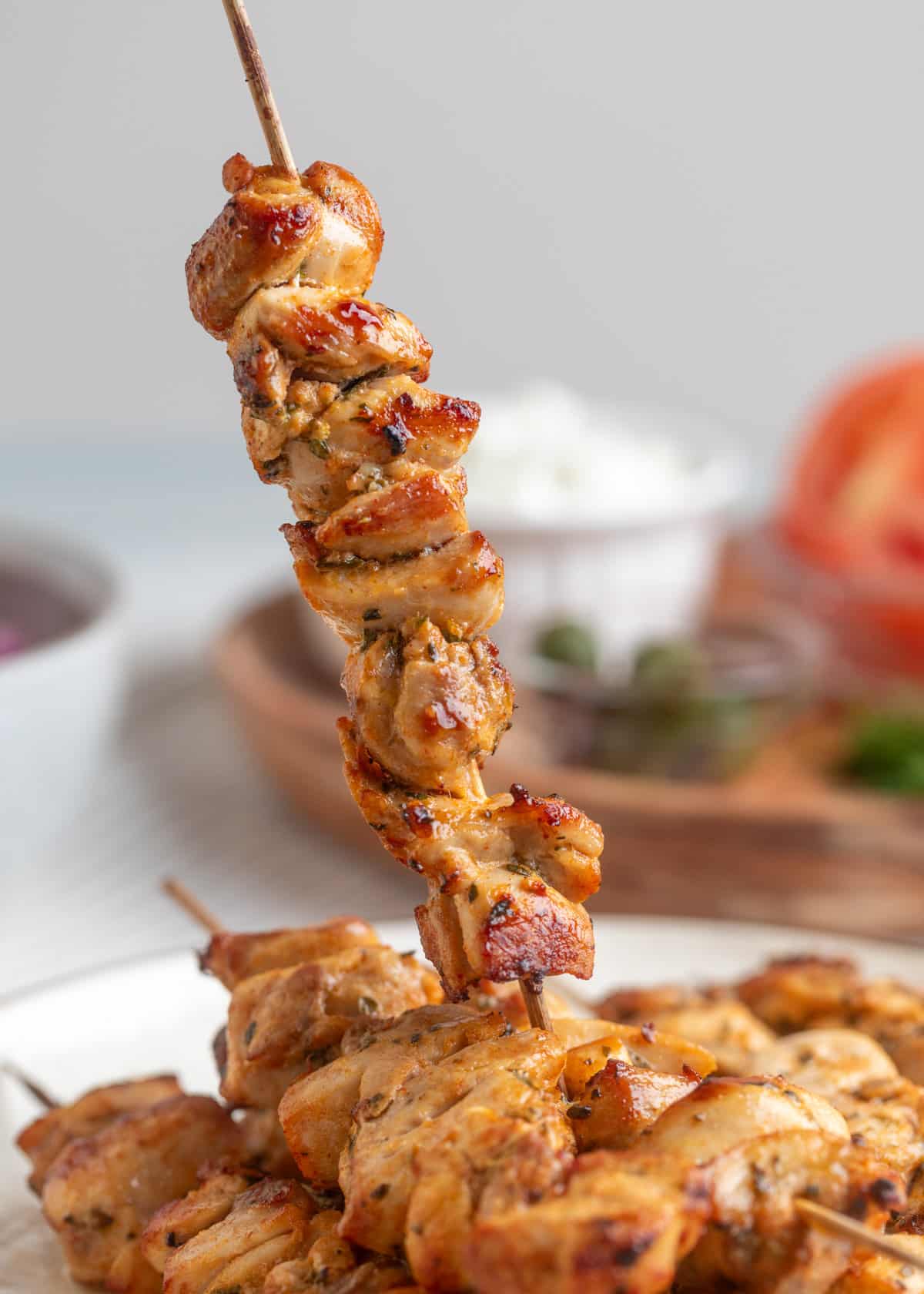 Close-up of a skewer of cooked chicken souvlaki being lifted from a plate, showing golden brown, juicy, and slightly charred pieces of marinated chicken. In the blurred background, small bowls of tzatziki, capers, and sliced tomato are visible.