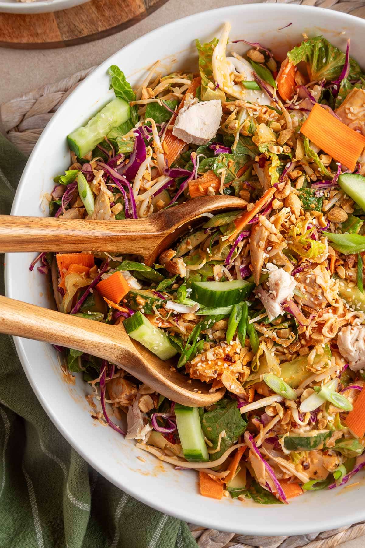 Overhead photo of Bang Bang Chicken Salad in a white bowl with wooden serving utensils. The salad is a colorful mix of shredded cabbage, romaine, cucumbers, carrots, chicken, green onions, chopped peanuts, and sesame seeds tossed in a creamy orange dressing, with a green striped napkin styled underneath.