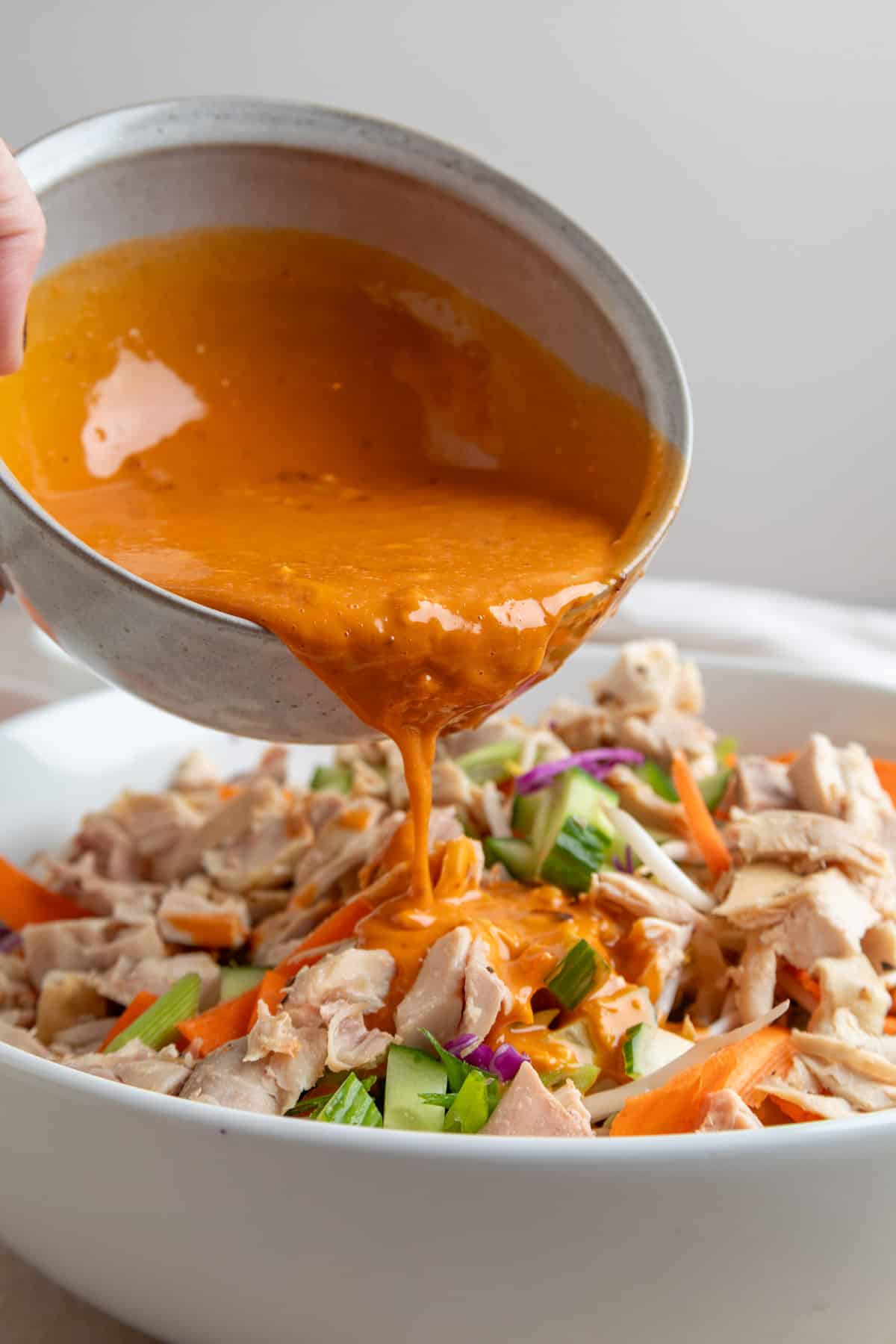 Close-up of a hand pouring creamy, orange Bang Bang sauce from a ceramic bowl over a salad made with chopped chicken, cucumbers, shredded cabbage, and carrot ribbons in a white serving bowl.