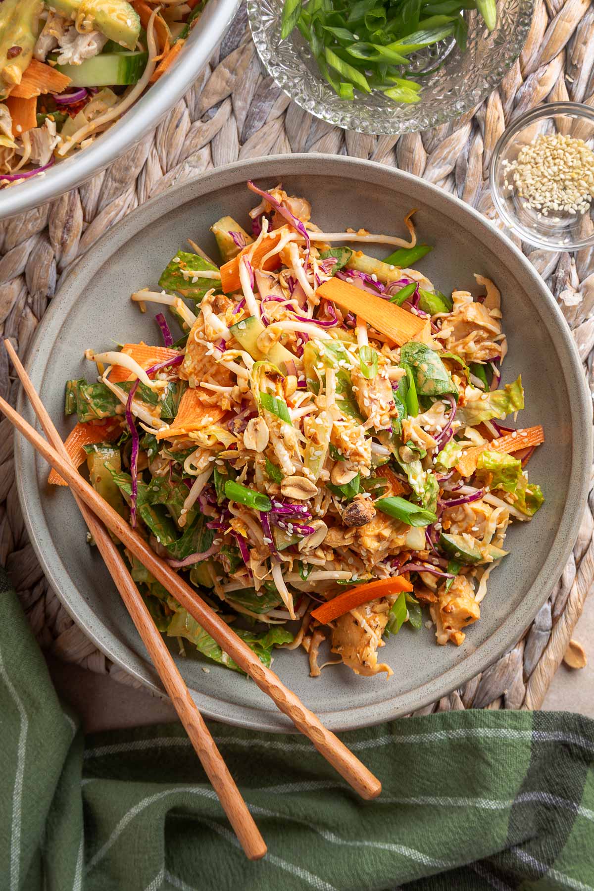 Overhead view of Bang Bang Chicken Salad served on a taupe ceramic plate with chopsticks. The salad features colorful shredded vegetables, chopped chicken, green onions, sesame seeds, and crushed peanuts on a woven placemat, with additional garnishes styled nearby.