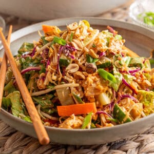 Side view of Bang Bang Chicken Salad served in a stoneware bowl with chopsticks, showcasing vibrant layers of shredded chicken, cabbage, carrots, cucumbers, green onions, and peanuts, all coated in a creamy, spicy dressing and sprinkled with sesame seeds.