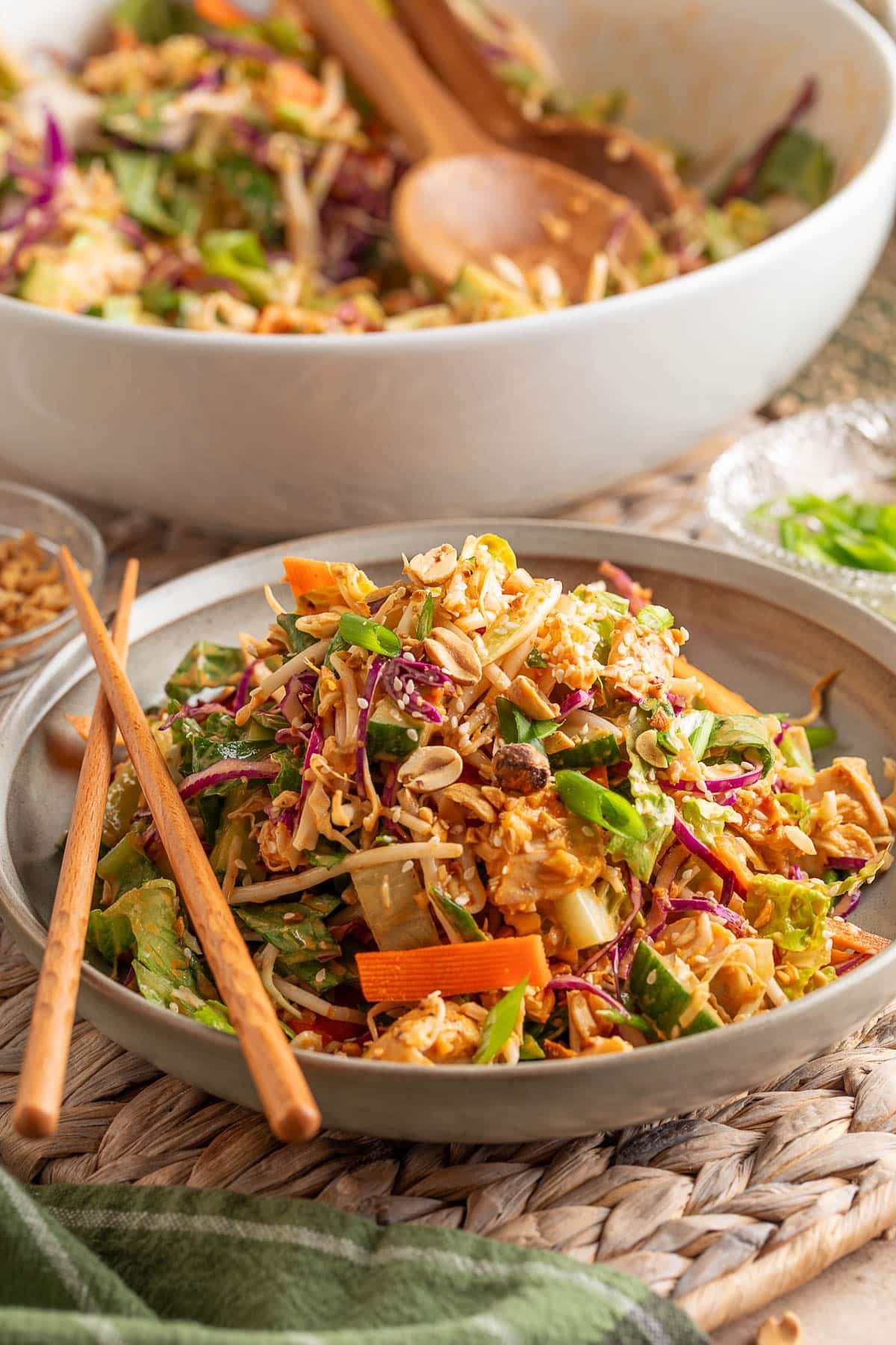Plate of Bang Bang Chicken Salad topped with peanuts, sesame seeds, and scallions, featuring shredded romaine, cabbage, carrots, cucumbers, and bean sprouts, with chopsticks resting on the side and a large serving bowl in the background.