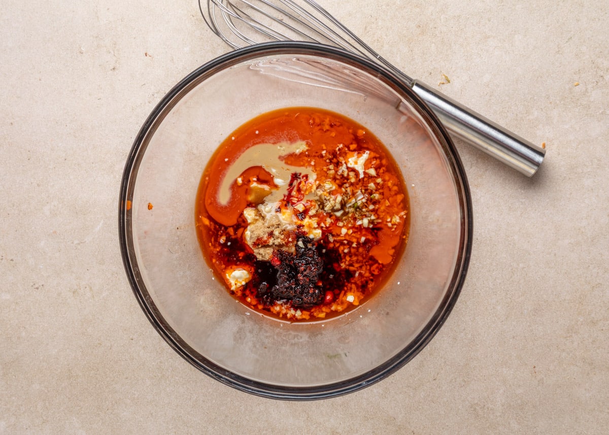 Unmixed bang bang dressing ingredients in a glass bowl, including chili oil, chili crunch, tahini, garlic, mayo, soy sauce, brown sugar, and ginger, with a metal whisk resting beside the bowl.