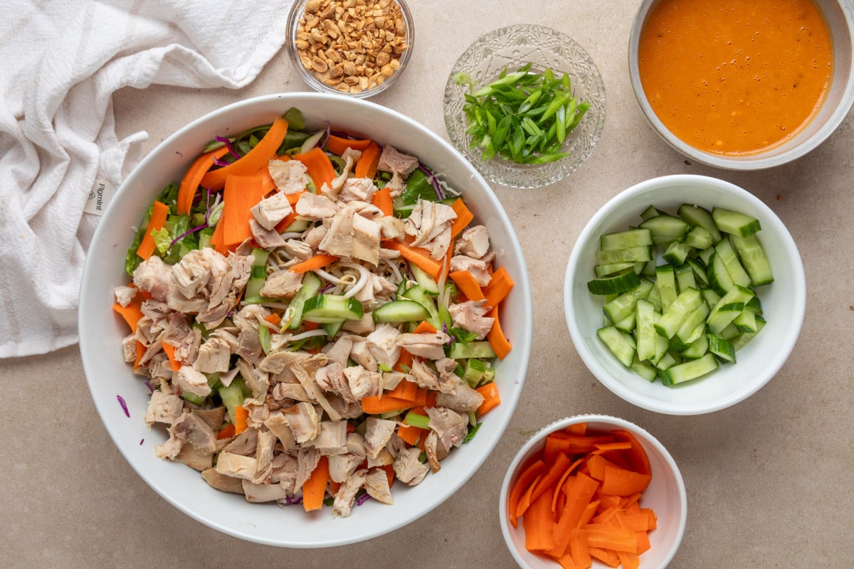 Overhead view of a large white bowl filled with chopped cooked chicken, romaine, shredded cabbage, cucumbers, carrots, and bean sprouts for Bang Bang Chicken Salad. Surrounding the bowl are small dishes with additional salad ingredients: chopped peanuts, sliced scallions, creamy orange dressing, extra cucumbers, and carrot ribbons. A white kitchen towel is partially visible in the corner.
