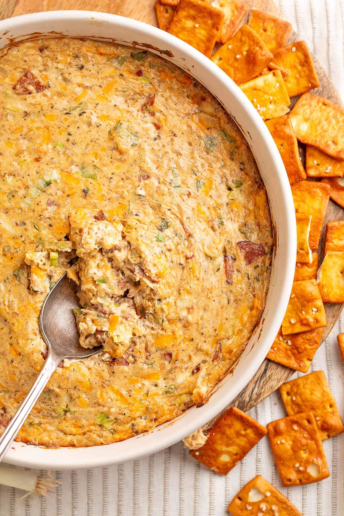 Overhead view of a warm, cheesy boudin dip in a white baking dish with a spoonful scooped from the center, surrounded by square buttery crackers on a wooden board.