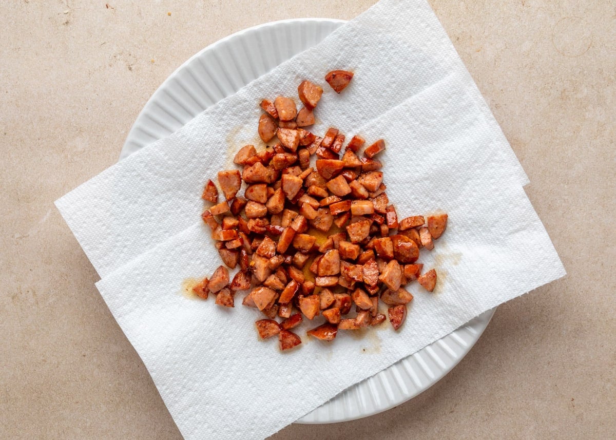 Cooked andouille sausage pieces draining on a white paper towel-lined plate, with glistening oil spots visible around the meat.