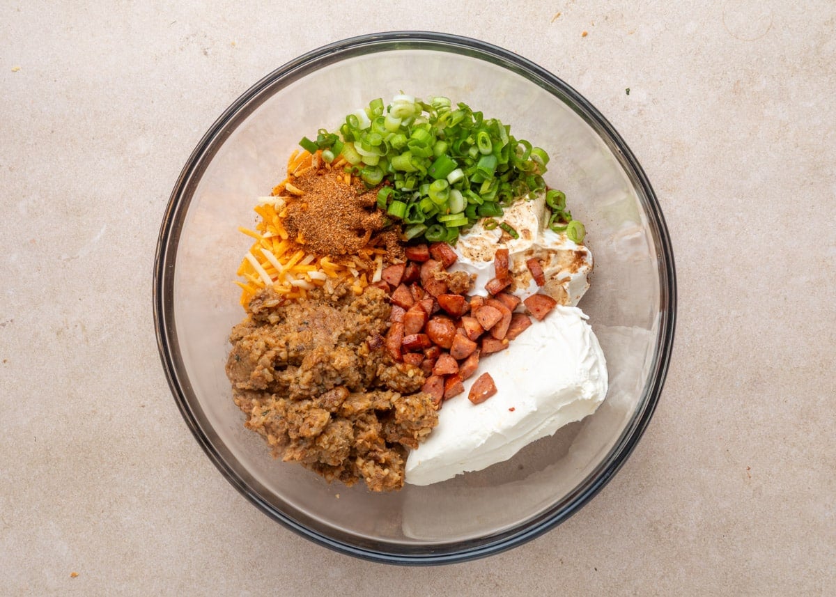 Top-down view of a glass mixing bowl with separated ingredients for Boudin Dip, including cream cheese, chopped cooked sausage, shredded cheese, sliced green onions, Creole seasoning, boudin filling, and sour cream.