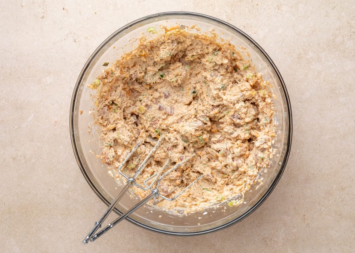 Top-down view of fully combined Boudin Dip in a glass mixing bowl with electric mixer beaters resting inside the creamy, speckled mixture.