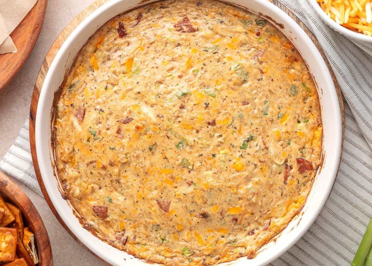 Close-up of a round white baking dish filled with golden-brown baked boudin dip, dotted with melted cheese, herbs, and pieces of sausage, set on a striped kitchen towel with bowls of shredded cheese and dippers nearby.