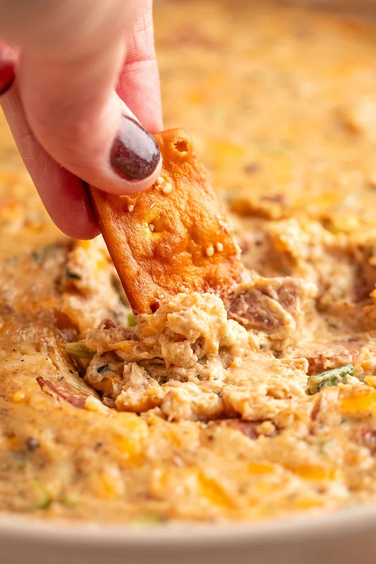 A hand with dark red nail polish dips a seasoned rectangular cracker into a hot, cheesy boudin dip filled with sausage, melted cheese, and green onions.