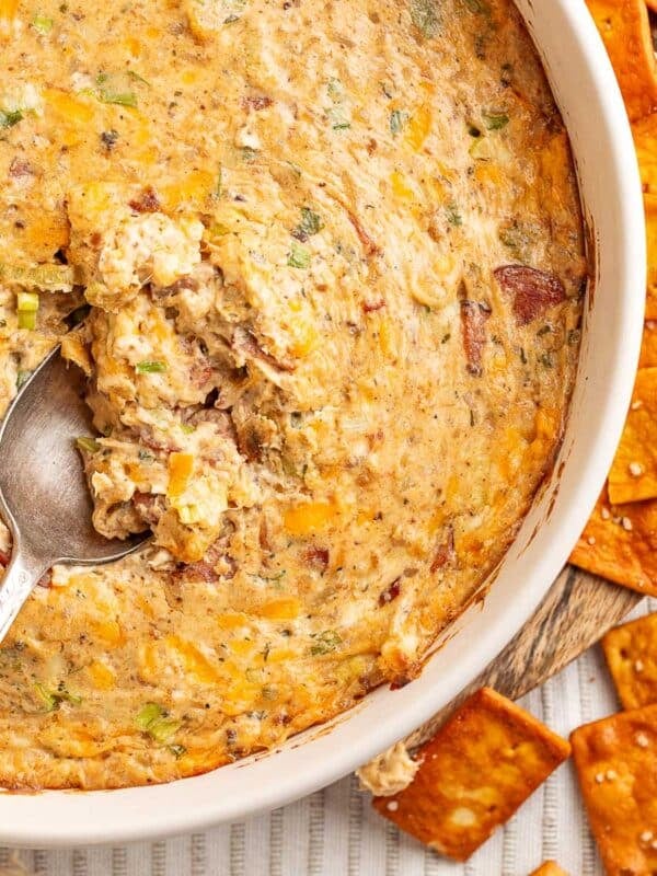 Close-up of Boudin Dip in a white baking dish with a serving spoon resting inside and crispy crackers scattered around the edge.
