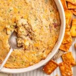 Close-up of Boudin Dip in a white baking dish with a serving spoon resting inside and crispy crackers scattered around the edge.
