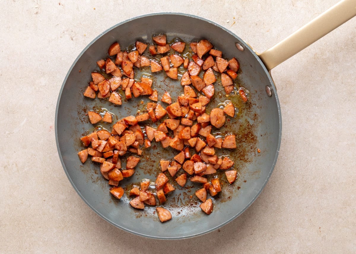 Chopped and browned andouille sausage pieces sizzling in a nonstick skillet, glistening with rendered fat and spices as they begin to caramelize.