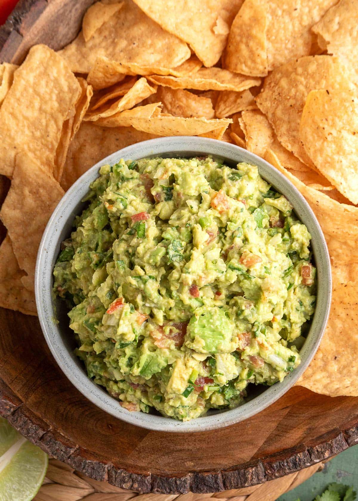 Overhead view of a rustic bowl filled with chunky guacamole made with avocado, tomatoes, onions, and cilantro, surrounded by golden tortilla chips on a wooden serving tray.