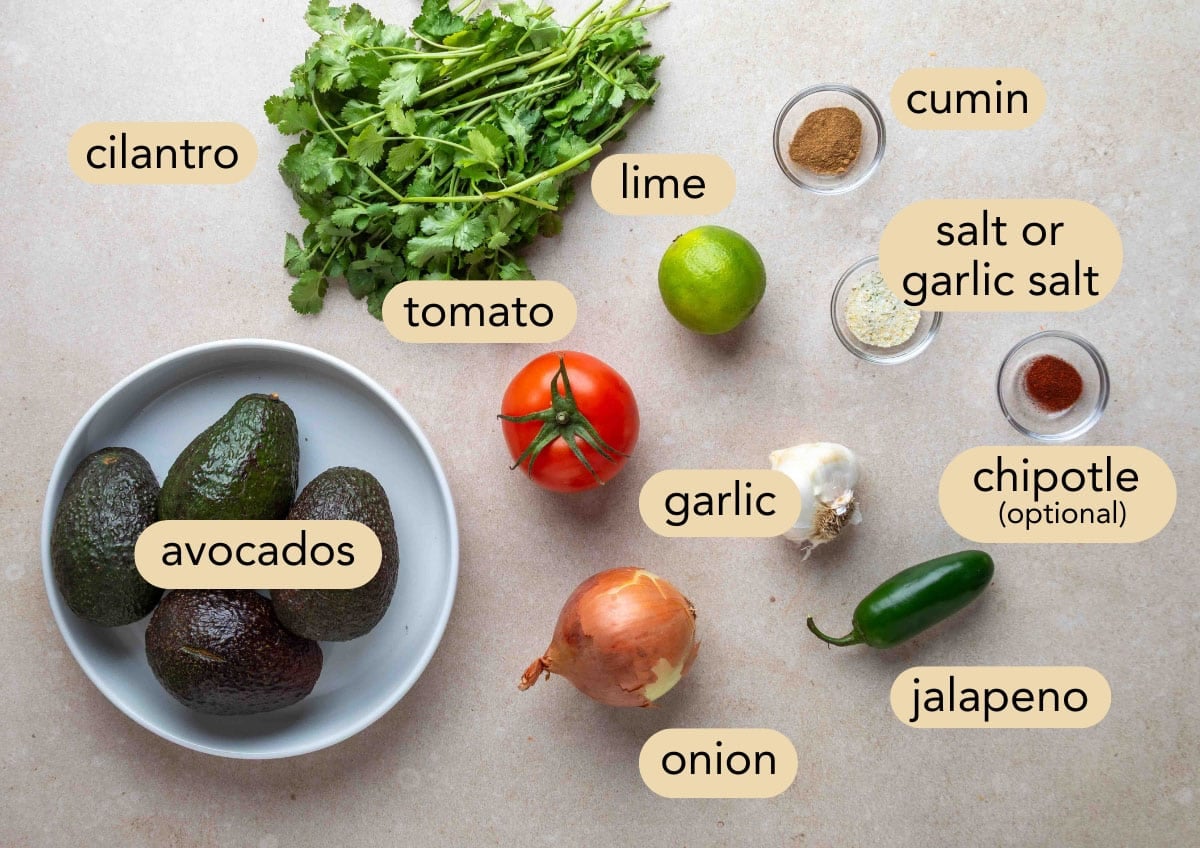 Labeled overhead photo of chunky guacamole ingredients arranged on a light surface, including avocados, tomato, onion, garlic, jalape&ntilde;o, cilantro, lime, cumin, chipotle powder (optional), and salt or garlic salt.