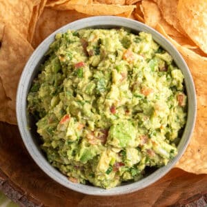 Overhead close-up of a rustic gray bowl filled with chunky guacamole, surrounded by tortilla chips on a wooden serving tray.