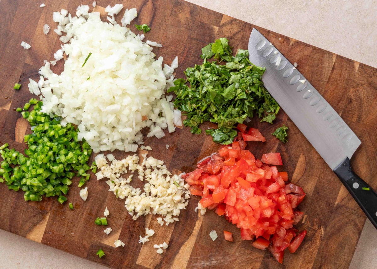 Chopped ingredients for guacamole&mdash;yellow onion, cilantro, tomato, garlic, and jalape&ntilde;o&mdash;arranged on a wooden cutting board next to a chef&rsquo;s knife.