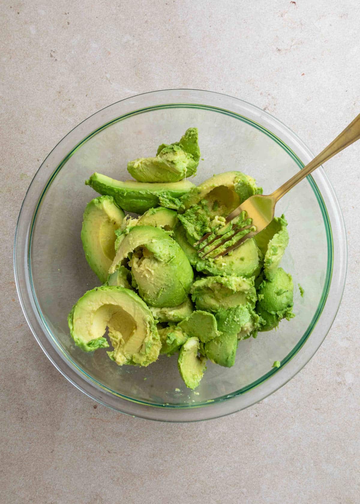 Halved and sliced avocados in a glass mixing bowl with a gold fork, ready to be mashed on a neutral countertop.